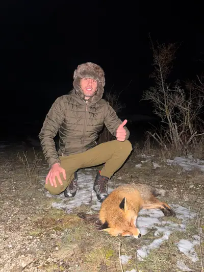 A hunter in a fur hat kneeling in thin snow at night beside a red fox, bare scrub behind