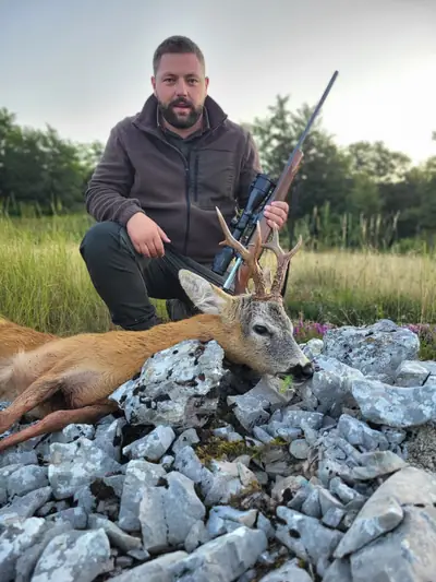 A hunter in a brown fleece kneeling with a rifle beside a roe buck on broken limestone