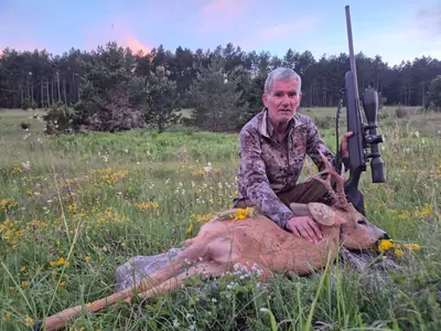 A hunter kneeling beside a roe buck in a flowering meadow in front of a pine wood at dusk