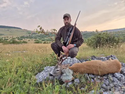 A hunter kneeling with a rifle beside a roe buck on the open plateau under a pink sunset sky