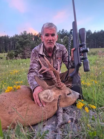 A hunter with a scoped rifle beside a roe buck among yellow wildflowers at dusk