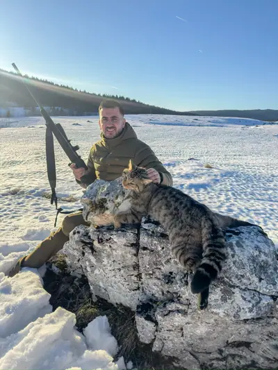 A hunter with a rifle sitting beside a wild cat laid on a limestone boulder, snow-covered plateau and forest ridge behind