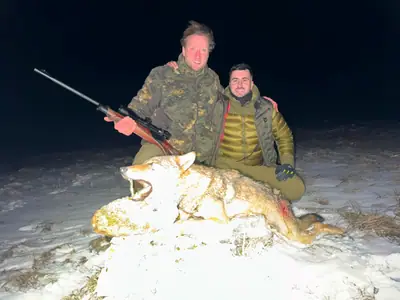Two hunters sitting together in the snow at night beside a wolf, smiling at the camera